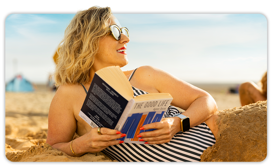 Close-up image of a woman in a blue swimming costume lying on the beach reading a book titled the good life 
